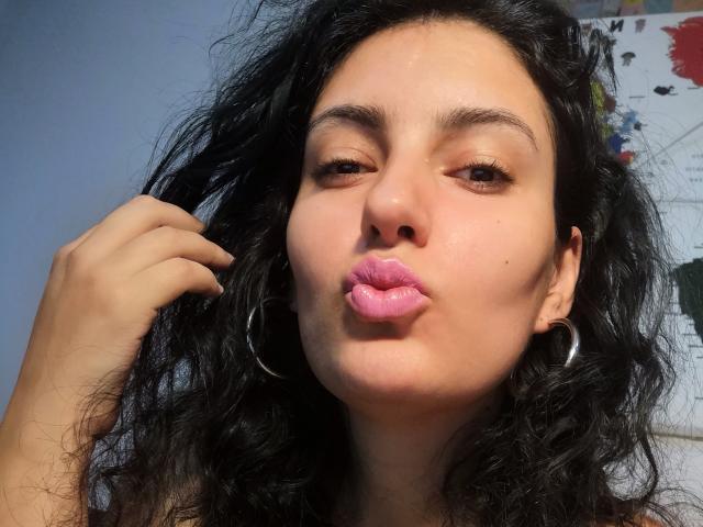 AddictedToUTS has curly black hair, pink lipstick, and is pouting with eyes half-closed, holding a lock of hair, wearing a silver hoop earring.