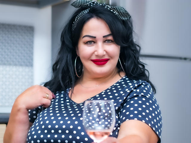 LexyRose with curly black hair, polka dot dress, red lipstick, hoop earrings, black bow, holding a glass, smiling, showing her big tits, in a kitchen.