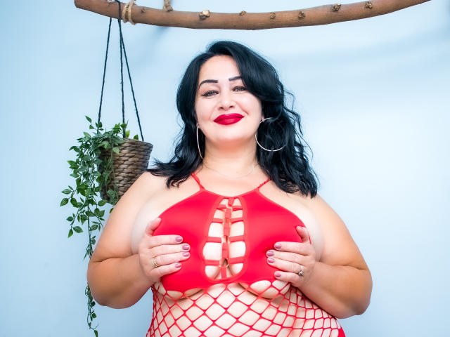 LexyRose with black wavy hair, red lipstick, and hoop earrings, wearing a red mesh top, covering her big boobs with her hands. Potted plant in background.