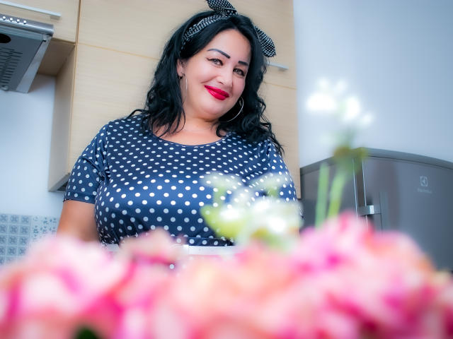 LexyRose, curvilínea com seios grandes, vestindo uma blusa de bolinhas, uma faixa de cabeça preta e batom vermelho, sorri em uma cozinha com flores rosa em primeiro plano..