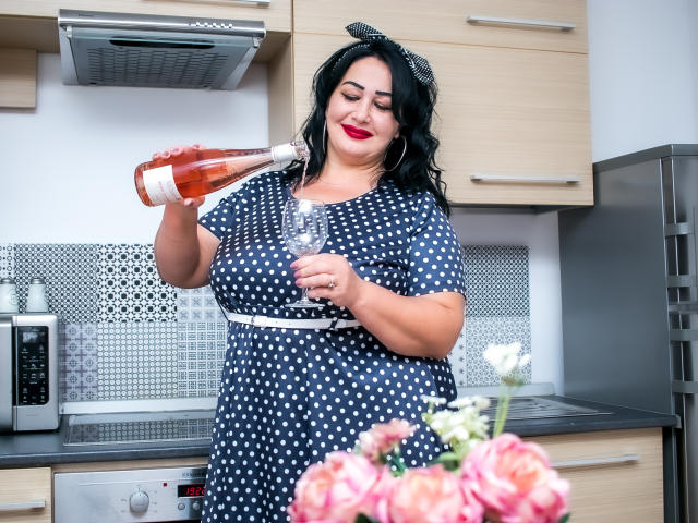 LexyRose with black hair, red lipstick, and polka dot dress pours wine into a glass, showing her curvy tits, in a kitchen with pink roses in the foreground.