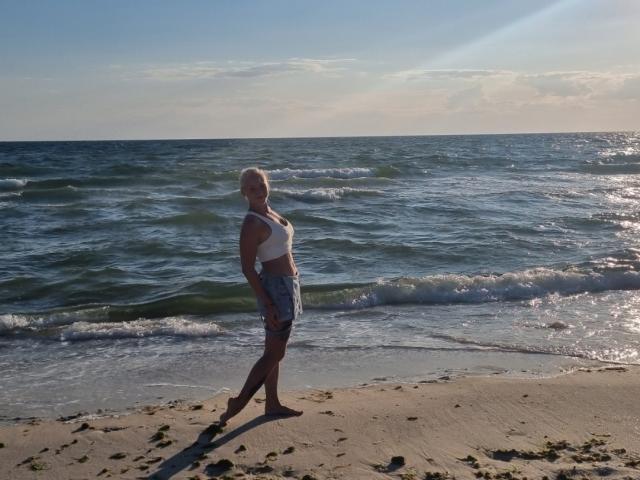 EllenSoNice stands on a beach, wearing a white top and blue shorts, with her back to the camera, her blonde hair blowing in the ocean breeze.