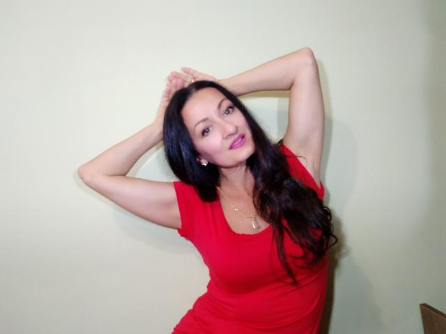 Antralyn, con cabello negro largo, vestido rojo, brazos levantados, manos detrás de la cabeza, sonriendo, lápiz labial rosa, piel clara, aretes pequeños, pared verde suave, ligera sombra, pose relajada..