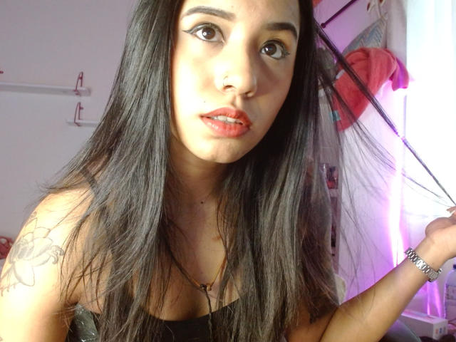 AshleyLover has long black hair, brown eyes, red lipstick, a nose piercing, and a tattoo on her left shoulder. She's wearing a black top and a silver bracelet.