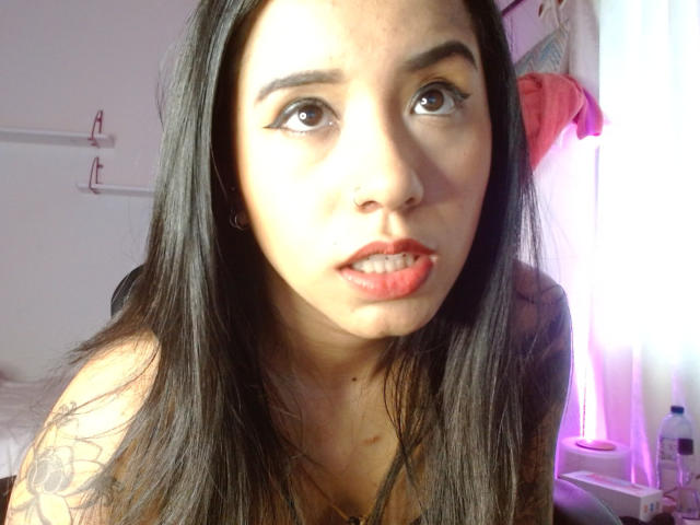 AshleyLover has long black hair, brown eyes, red lipstick, and a tattoo on her right arm. She's looking up with a slightly open mouth, showing her teeth.