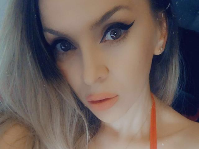 SophieDeee has blue eyes with thick black eyeliner, full pink lips, and blonde hair. She's wearing a red halter top, showing her neck and earrings.
