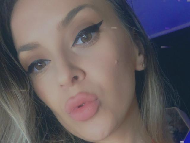 SophieDeee's face with pouty pink lips, thick black eyeliner, brown eyes, and blonde hair. She's wearing small earrings and has a slight pimple on her right cheek.