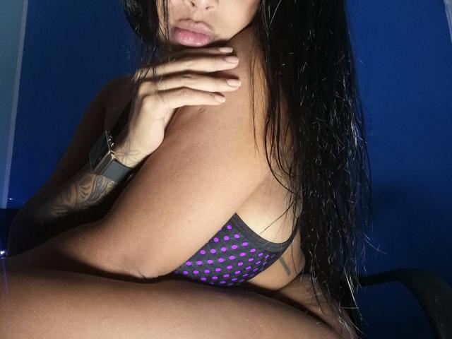 VickyGreys with wet black hair, purple polka dot bra, tattooed arm, wrist bracelet, hand on neck, partially visible large tits, dark blue background, and a subtle pout.