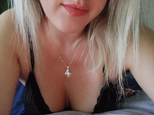 SweetDeliaa, com cabelo loiro, vestindo uma blusa preta de renda, mostrando os seios, um colar de ouro com um pingente de borboleta e um leve sorriso, inclinada para frente..