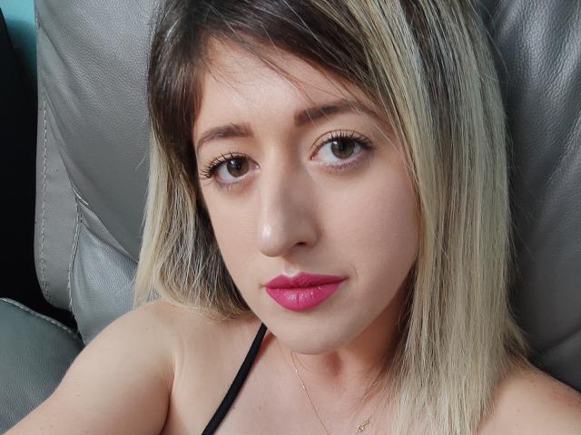 SweetDeliaa, con cabello rubio y castaño, lápiz labial rosa, ojos verdes, un sujetador negro y un maquillaje sutil, mirando a la cámara, sentada en una silla de cuero gris..
