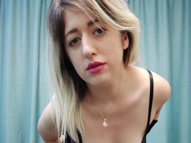 A SweetDeliaa tem cabelo loiro, na altura dos ombros, batom rosa e um colar de ouro com um pequeno pingente, está inclinada para frente, vestindo uma blusa preta, mostrando seus pequenos seios..