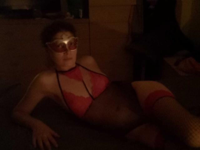 Kaylay è sdraiata su un letto, indossa un intimo di pizzo rosso con inserti in rete nera, calze rosse alte fino alla coscia e occhiali da sole bianchi. Ha il seno piccolo e un sedere scuro..