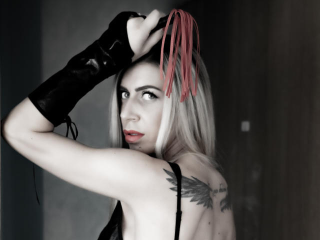 AnnesWhite with pale skin, blonde hair, red fangs in hair, black glove, wing tattoo on back, looking over shoulder, red lipstick, gray background.