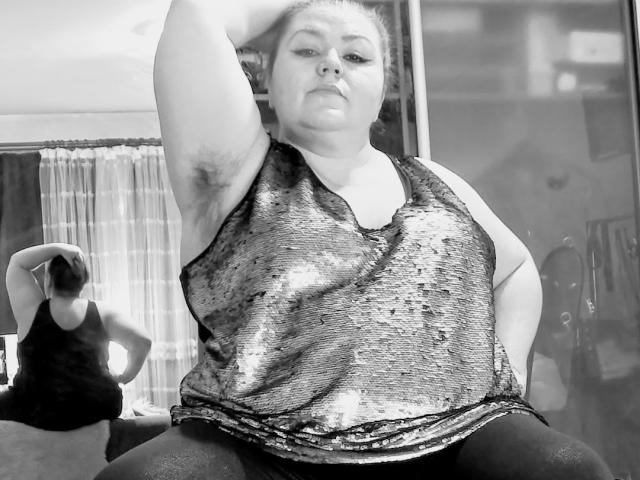 KaryQueen ist eine mollige, übergewichtige Frau mit Achselhaaren, die ein glänzendes, pailletbestreutes Top ohne Ärmel trägt und mit einem Arm erhoben sitzt, wobei ihre Achsel zu sehen ist. Spiegelbild im Hintergrund..