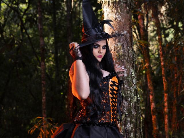 JassmineTS con bustino nero con lacci, cappello da strega, guanti a rete, e lunghi capelli neri, in piedi vicino a un albero, sfondo di foresta, con il trucco, che guarda la fotocamera, tenendo in mano il cappello..