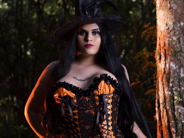 JassmineTS mit langen, schwarzen Haaren, trägt ein orange-schwarzes Schnürcorsett, einen schwarzen Hut mit Federn und hat Tattoos auf der Brust. Sie steht vor einem Baum, im Hintergrund ist ein Wald zu sehen..