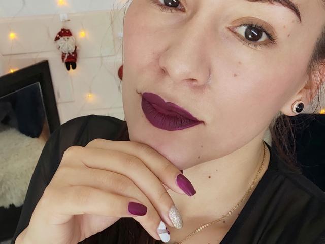 Maloryxx has fair skin, dark purple lipstick, and matching nail polish. She's wearing a black top, gold necklace, and black ear gauges, with a cute gnome decoration in the background.