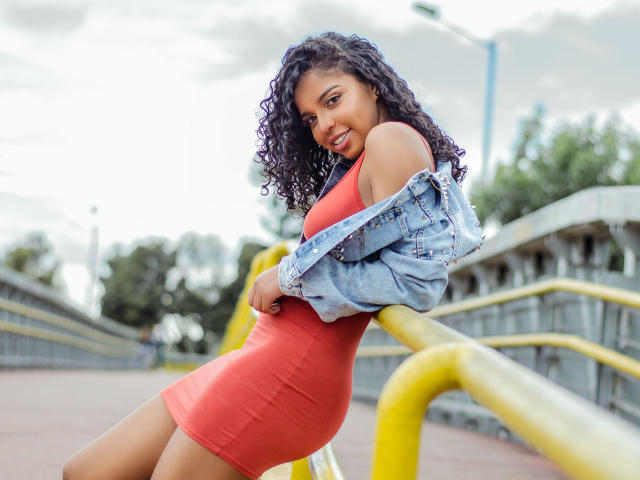 LisaMendez with curly black hair, wearing a tight red dress, and a denim jacket, leaning on yellow railings, smiling, showing her ass and a cute face.