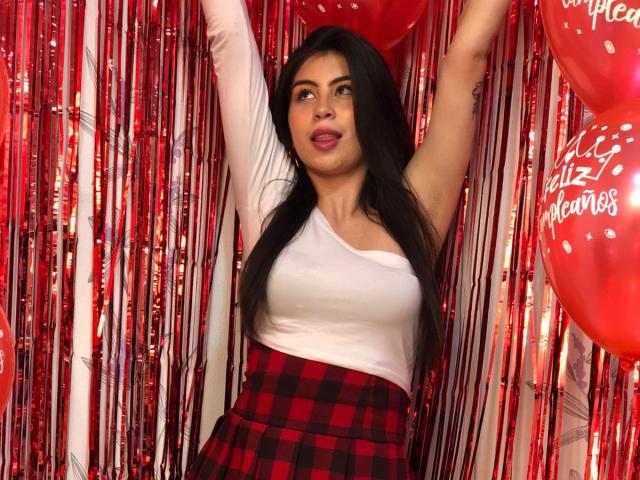 IsaTimeSweet has long black hair, a white one-shoulder top, and a red checkered skirt, raising arms, with a confident, slightly open-mouthed expression. Red balloons and streamers.