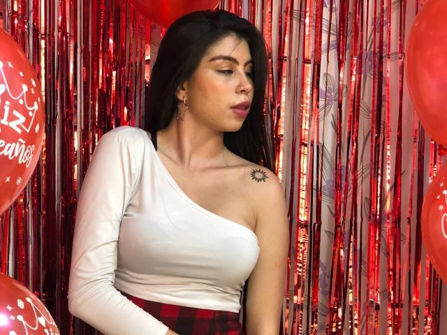 IsaTimeSweet, con i capelli scuri, indossa una maglietta bianca asimmetrica e una gonna a quadri rossa, mostrando un tatuaggio del sole sulla spalla destra, circondata da palloncini rossi, e guarda di lato..