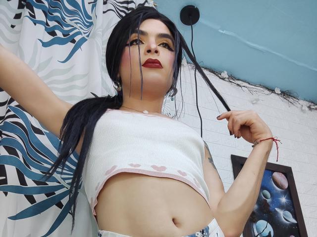 Rosariio with black pigtails, red lipstick, white crop top showing her tits, and blue jeans, standing in a room with a zebra-patterned curtain. Her right arm is raised.