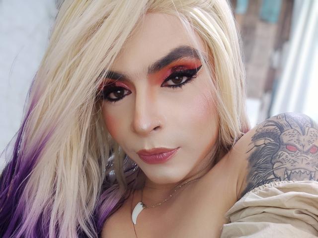 Rosariio hat platinblondes und violettes Haar, auffälliges rotes und schwarzes Augen-Make-up, eine tätowierte Schulter und trägt eine weiße Muschelkette mit rosafarbenen Lippen und einem leichten Lächeln..