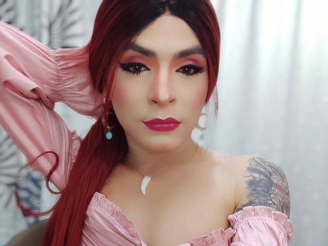 Rosariio with red hair, pink off-shoulder top, pink eyeshadow, white teardrop earrings, pink lipstick, small tattoo on right shoulder, hand in hair, looking at camera.
