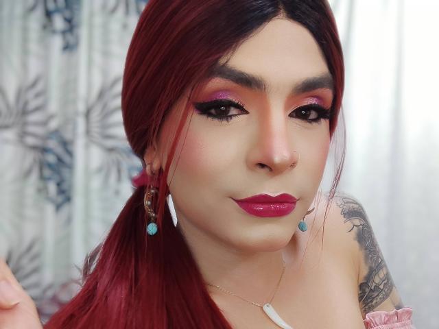 Rosariio mit roten Haaren, rosa Lidschatten, dunklem Eyeliner, glänzenden rosa Lippen, Nasenring, blauen Ohrringen, Tattoo an der rechten Schulter, hält die Haare in der linken Hand..
