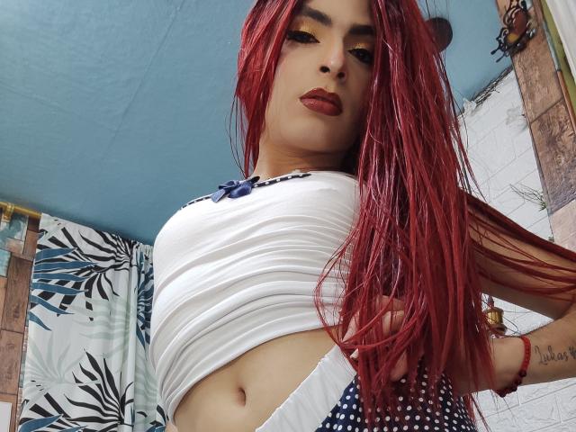 Rosariio met rood haar, een witte crop top en een rok met stippen, die haar borsten, kleine billen en rode lippenstift laat zien, en die naar beneden kijkt naar de camera..