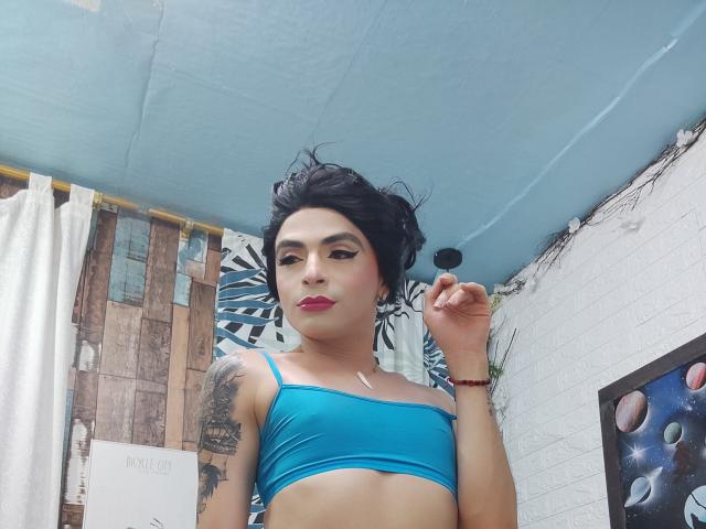 Rosariio met kort zwart haar, een blauw topje, een tatoeage op haar rechterarm, rode lippenstift, die een zwarte haarspeld vasthoudt, staat in een kamer met witte en blauwe muren..