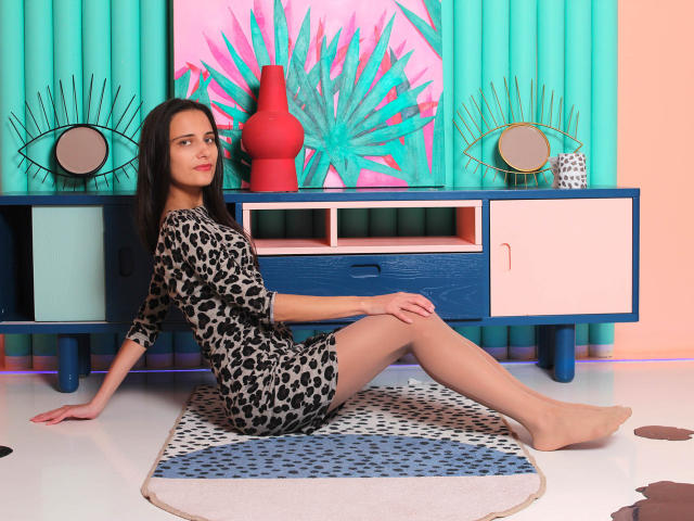 Orfeya, sitting on a patterned rug, wearing a leopard-print dress, has a slender build, small tits, and is looking at the camera with a slight smile.