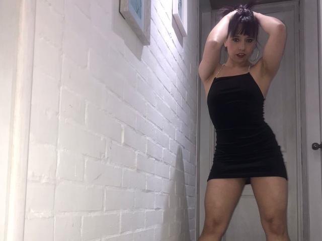 BellaLolita se tient dans un couloir, vêtue d'une mini-robe noire moulante, les mains dans ses cheveux foncés, mettant en valeur ses bras et ses jambes musclés, ainsi que ses fesses..