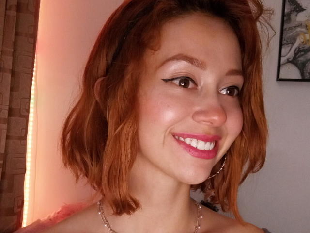 Eliza Jones, avec des cheveux roux, un eye-liner noir, du rouge à lèvres rose, souriante, portant un collier, des boucles d'oreilles créoles, une peau claire, et avec des fesses visibles en arrière-plan..