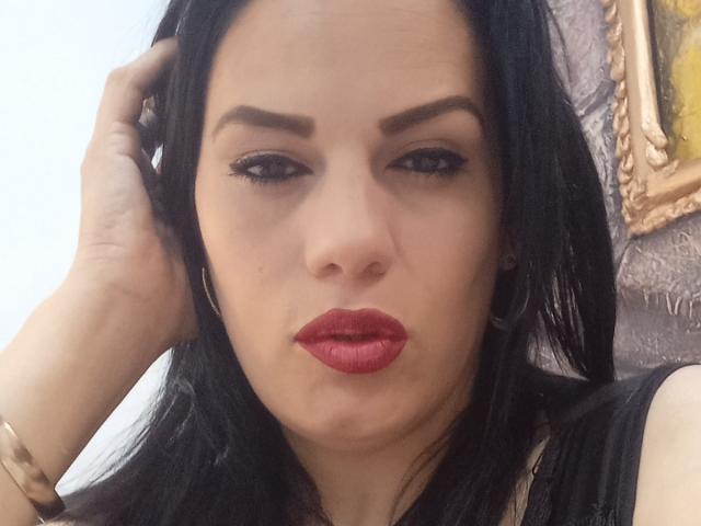 LizaRussell has dark black hair, red lipstick, thick eyebrows, and a serious expression. She's wearing a black top and a gold bracelet, with a hand in her hair.