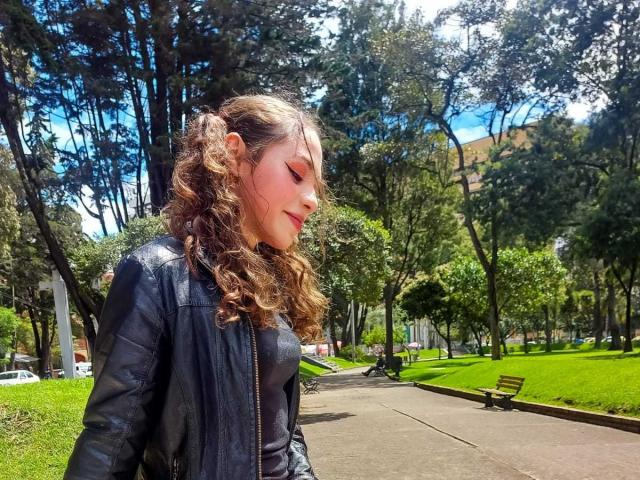 ArianaaMoon, con cabello castaño rizado, vestida con una chaqueta de cuero negra, de pie en un parque, con los ojos cerrados, la luz del sol en su rostro, un camino de hierba y un banco de madera al fondo..
