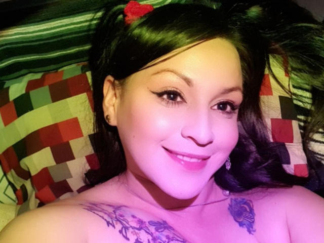 ElinaJhonson has dark hair, a red hairclip, and a smile showing her white teeth. She's topless with tattoos on her chest and wearing small earrings.