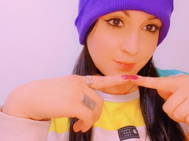 ElinaJhonson with a purple beanie, black eyeliner, pink lips, and long dark hair, pointing at a ring with pink nail polish, wearing a yellow and white shirt.