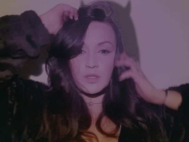 ElinaJhonson has wavy dark brown hair, her hand in it, wearing a black fuzzy jacket, light pink skin, subtle makeup, and a necklace, looking slightly to the side.