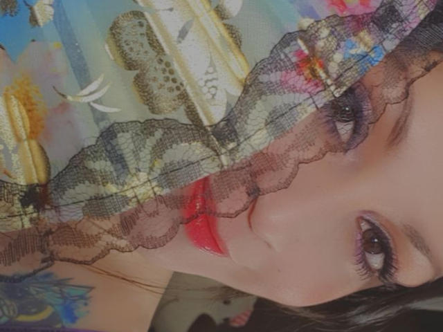 ElinaJhonson's face with red lips, black lace over her eyes, and a colorful floral dress, her brown eyes looking forward. She's got a sexy, cute look.