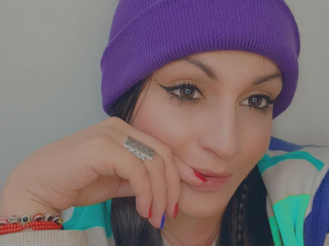 ElinaJhonson with purple beanie, black eyeliner, red nails, silver ring, and blue eyes, resting her chin on her hand, wearing a green and blue striped shirt.