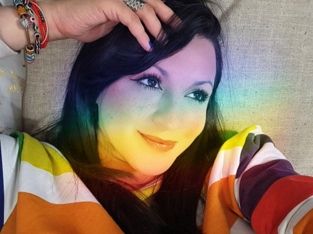 O rosto de ElinaJhonson está pintado com um degradê de cores do arco-íris, ela veste uma camisa colorida listrada, está deitada em uma superfície bege, com uma mão tocando sua cabeça, mostrando vários braceletes..