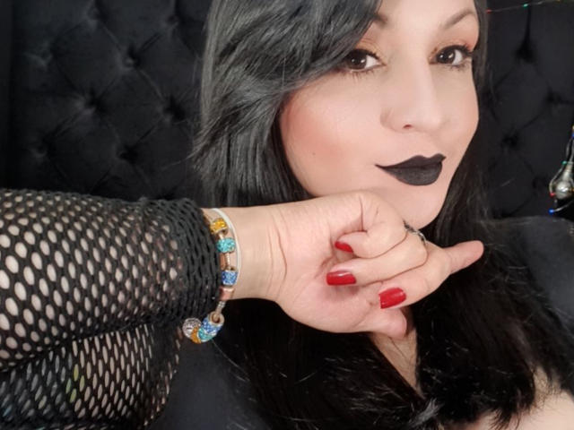 ElinaJhonson with black lipstick, red nails, fishnet sleeve, and colorful bracelet, holding her hand to her mouth, black hair, dark eyes, and tufted black headboard in background.