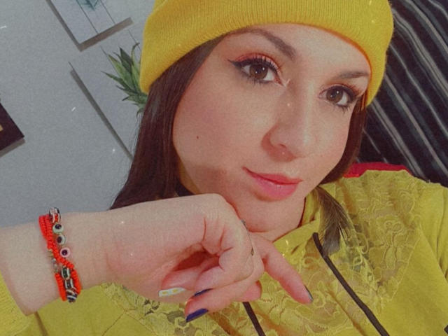 Elina Jhonson, con un gorro amarillo, ojos marrones y delineador de ojos rosa, vestida con una camisa amarilla con cordones, pulseras rojas y naranjas, y uñas azules, tocándose la barbilla..