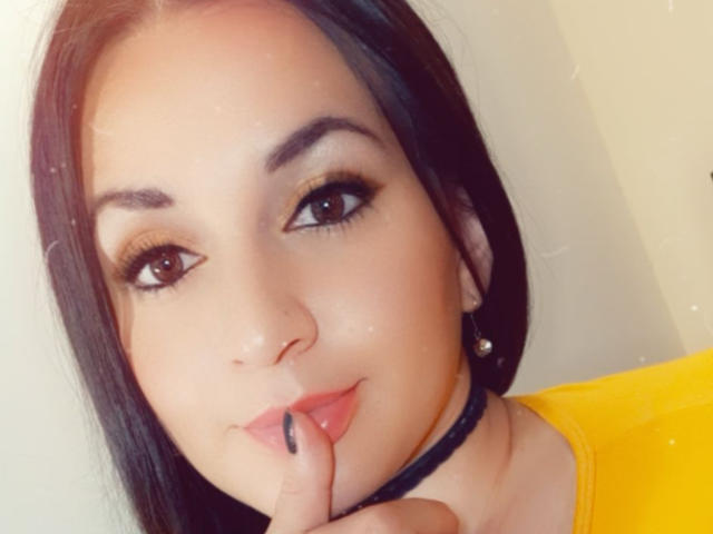 ElinaJhonson tem um rosto bonito, cabelo preto, olhos castanhos e um nariz pequeno, com um dedo nos lábios. Ela está vestindo uma camisa amarela e um colar preto..