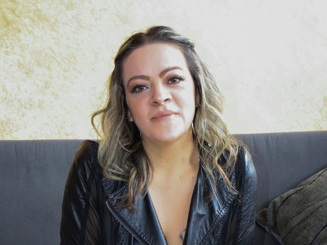 LiliCarina hat welliges, blondes Haar, helle Haut und trägt eine schwarze Lederjacke. Sie sitzt auf einem grauen Sofa, mit einem leichten Lächeln und einem entspannten Gesichtsausdruck..