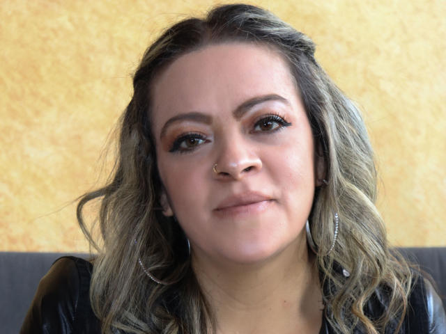 LiliCarina tiene senos de tamaño medio, un piercing en la nariz y cabello ondulado de color rubio ceniza. Lleva una blusa de cuero negro con aretes de plata, y tiene una sonrisa leve y segura..
