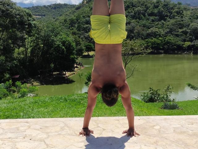 JeanChaud è a testa in giù, sta facendo la verticale su un patio di pietra, indossa dei pantaloncini gialli, sullo sfondo si vede una zona erbosa, uno stagno e delle colline boscose..