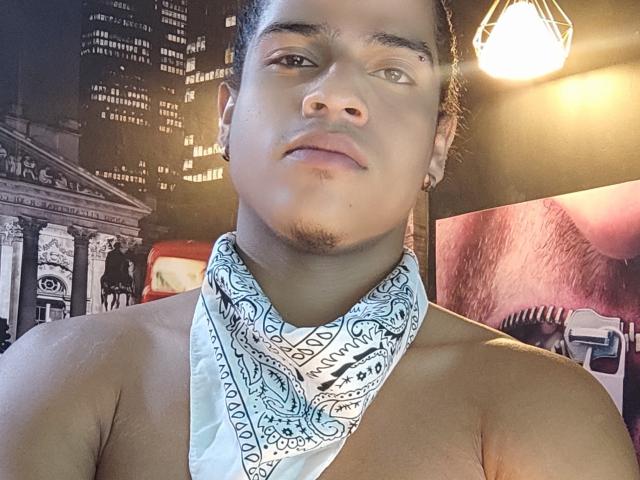 JeanChaud, avec une peau marron moyen, des cheveux noirs courts, une petite moustache, portant un bandana blanc, torse nu, sous une lumière suspendue, regardant légèrement sur le côté, avec une expression neutre..