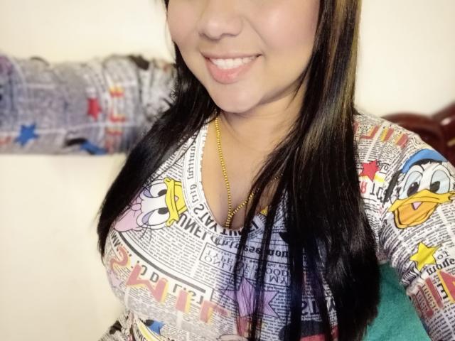 AvaSexySeins met lang zwart haar, lachend, gekleed in een kleurrijke top met een krantenpatroon met afbeeldingen van Donald Duck en Mickey Mouse, waarbij haar borsten zichtbaar zijn. Gouden ketting, witte achtergrond..