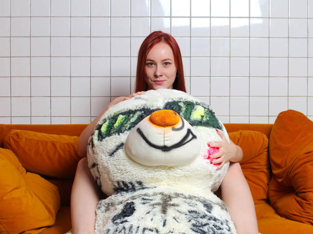 Lilian Kroft, con i capelli rossi, che tiene in braccio un enorme peluche a forma di pinguino, seduta su un divano arancione, con il suo grande sedere a malapena coperto dal pinguino, sorride alla telecamera..